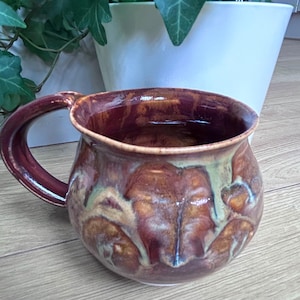 May include: A round ceramic mug featuring a glossy glaze with blended tones of deep burgundy, warm brown, and muted sage green. The mug has a wide, curved body and a thick, sturdy handle attached to the side, showcasing a smooth, reflective finish.