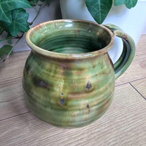 May include: A round ceramic mug with a glossy finish in shades of olive green and brown. The mug features a wide, bulbous body with horizontal ridges along the exterior surface, a flared rim, and a curved handle attached to the side of the vessel.