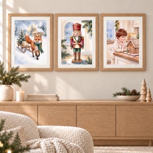 May include: Three framed watercolor prints displayed on a wall. The left print shows a fox in a green scarf pulling a sled with a gift. The center print features a red and green nutcracker. The right print depicts a child decorating a gingerbread house.