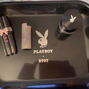May include: A black rolling tray with the Playboy logo and the text "PLAYBOY by RYOT". The tray holds a black lighter case with a pink bow, a rhinestone lighter, and a black grinder with the Playboy logo.