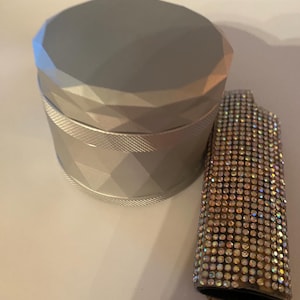 May include: A silver grinder with a faceted design and a rhinestone-covered lighter. The grinder is cylindrical with a textured band. The lighter is covered in iridescent rhinestones.