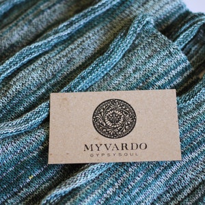 Puede incluir: Bufanda de punto doblada en tonos turquesa, blanco y gris. Una etiqueta rectangular con las palabras "MYVARDO GYPSYSOUL" y un diseño circular se coloca encima de la bufanda.
