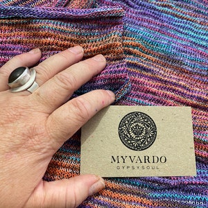 Op de afbeelding: Een veelkleurige gebreide sjaal in tinten paars, oranje en blauw. De sjaal wordt vastgehouden door een hand met een zilveren ring. Een klein, rechthoekig label met de woorden "MYVARDO GYPSYSOUL" is ook zichtbaar.