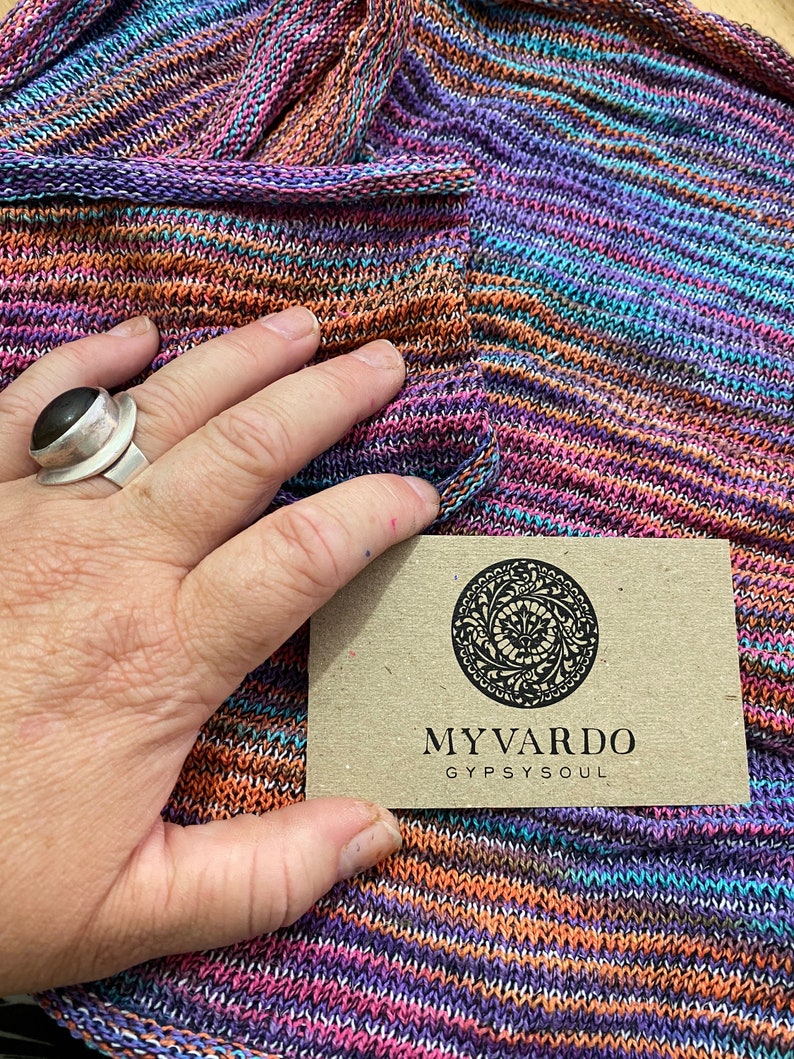 Op de afbeelding: Een veelkleurige gebreide sjaal in tinten paars, oranje en blauw. De sjaal wordt vastgehouden door een hand met een zilveren ring. Een klein, rechthoekig label met de woorden "MYVARDO GYPSYSOUL" is ook zichtbaar.
