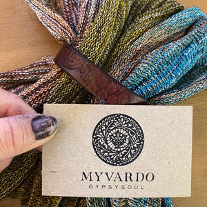 May include: A knitted scarf in various colours, including green, blue, orange, and brown, held together with a brown leather cuff. A business card with the text "MYVARDO GYPSYSOUL" and a circular design is in the foreground.