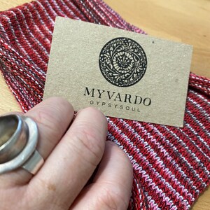 Op de afbeelding: Een close-up van een rood, wit en zwart gestreepte stof met een bruin label. Het label heeft een cirkelvormig ontwerp en de woorden "MYVARDO GYPSYSOUL". Een zilveren ring is zichtbaar.
