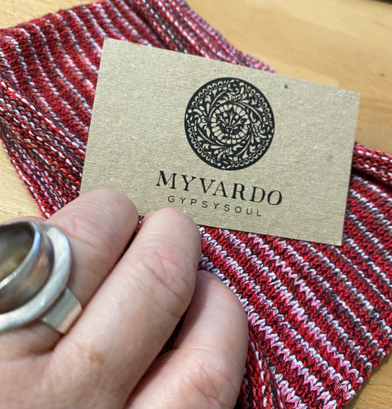 Op de afbeelding: Een close-up van een rood, wit en zwart gestreepte stof met een bruin label. Het label heeft een cirkelvormig ontwerp en de woorden "MYVARDO GYPSYSOUL". Een zilveren ring is zichtbaar.