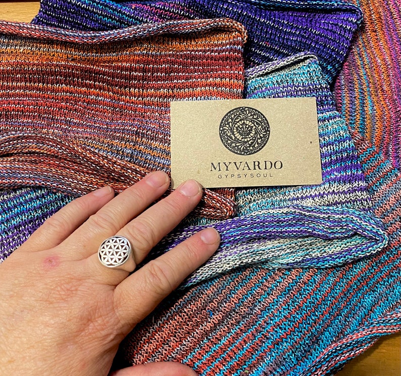 Op de afbeelding: Close-up van kleurrijke gebreide stof in tinten oranje, paars en blauw. Een zilveren ring met een geometrisch ontwerp zit aan een hand. Een kleine bruine kaart met de tekst "MYVARDO GYPSYSOUL" is ook zichtbaar.