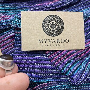 Op de afbeelding: Close-up van een gebreide stof in blauwe, paarse en roze tinten. Een kleine, bruine kaart met de tekst "MYVARDO GYPSYSOUL" ligt op de stof. Een zilveren ring is zichtbaar in de linker benedenhoek.