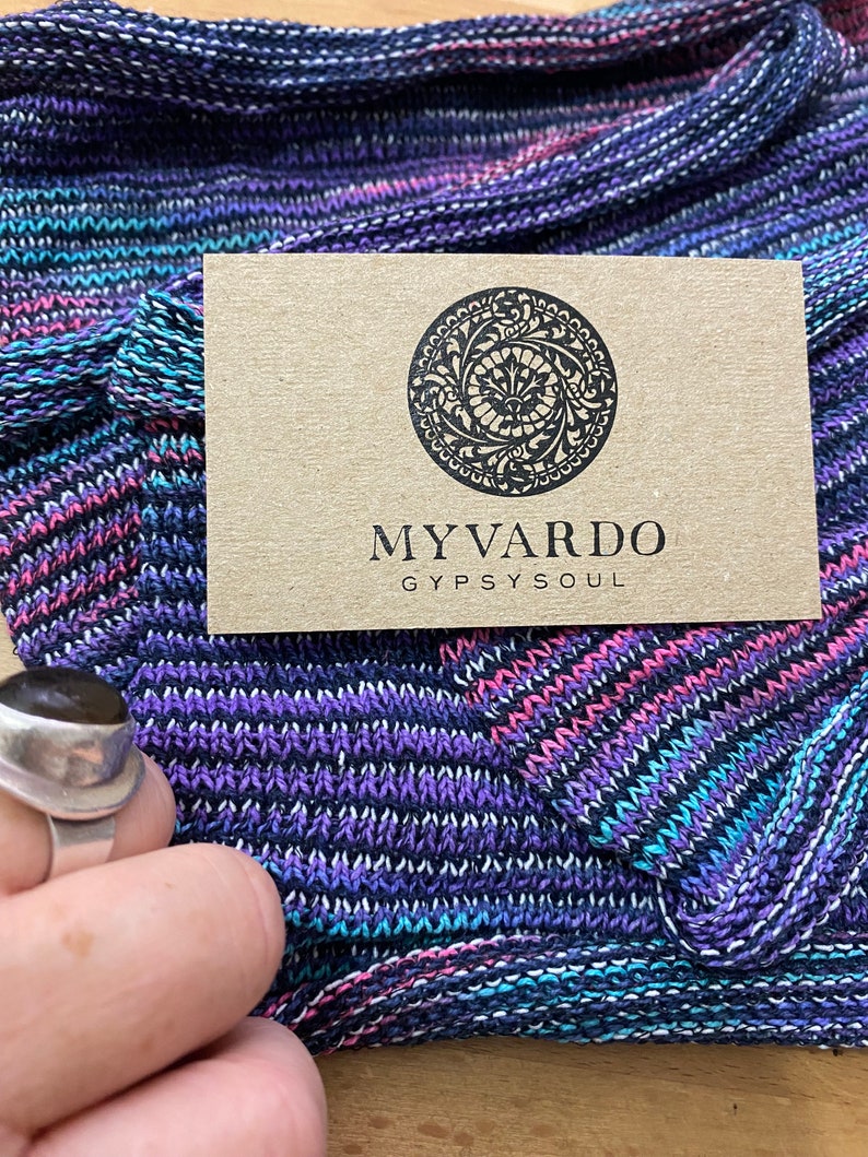 Op de afbeelding: Close-up van een gebreide stof in blauwe, paarse en roze tinten. Een kleine, bruine kaart met de tekst "MYVARDO GYPSYSOUL" ligt op de stof. Een zilveren ring is zichtbaar in de linker benedenhoek.