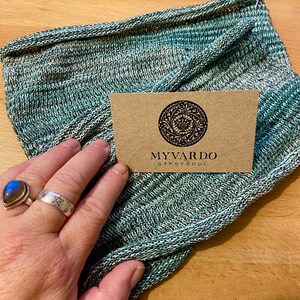 Op de afbeelding: Een groen, witte en zwarte gebreide sjaal met de tekst "MYVARDO / GYPSY SOUL" op een bruine kaart. De sjaal is opgevouwen en toont het textuurpatroon. Een hand met twee ringen is zichtbaar.