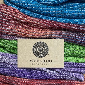 May include: A stack of colourful knitted scarves in shades of blue, red, green, purple, and brown. The scarves are made from a soft, textured yarn and have a subtle shimmer. A brown paper tag with the text "MYVARDO GYPSYSOUL" is visible in the centre of the image.