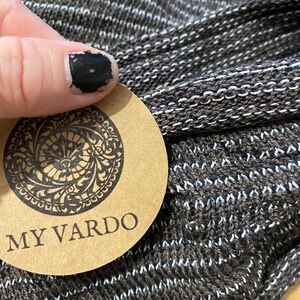 Op de afbeelding: Close-up van een bruine en witte gebreide stof. De stof heeft een textuur met horizontale witte strepen. Een rond label met de woorden "MY VARDO" is zichtbaar. Het label heeft een sierlijk ontwerp rond de rand.