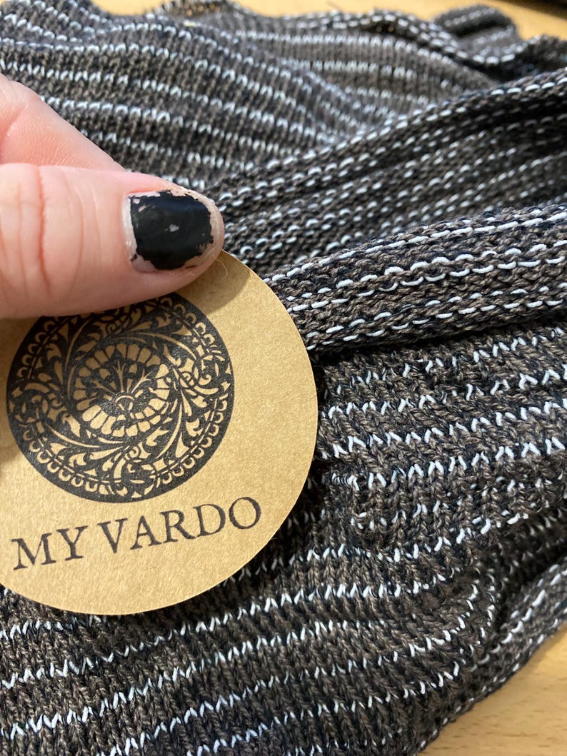 Op de afbeelding: Close-up van een bruine en witte gebreide stof. De stof heeft een textuur met horizontale witte strepen. Een rond label met de woorden "MY VARDO" is zichtbaar. Het label heeft een sierlijk ontwerp rond de rand.