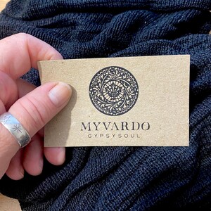 Op de afbeelding: Een lichtbruine visitekaart met de tekst "MYVARDO GYPSYSOUL" en een cirkelvormig ontwerp wordt boven een donkerblauwe, getextureerde stof gehouden. Een zilveren ring met sterontwerpen is zichtbaar. De stof lijkt een sjaal te zijn.