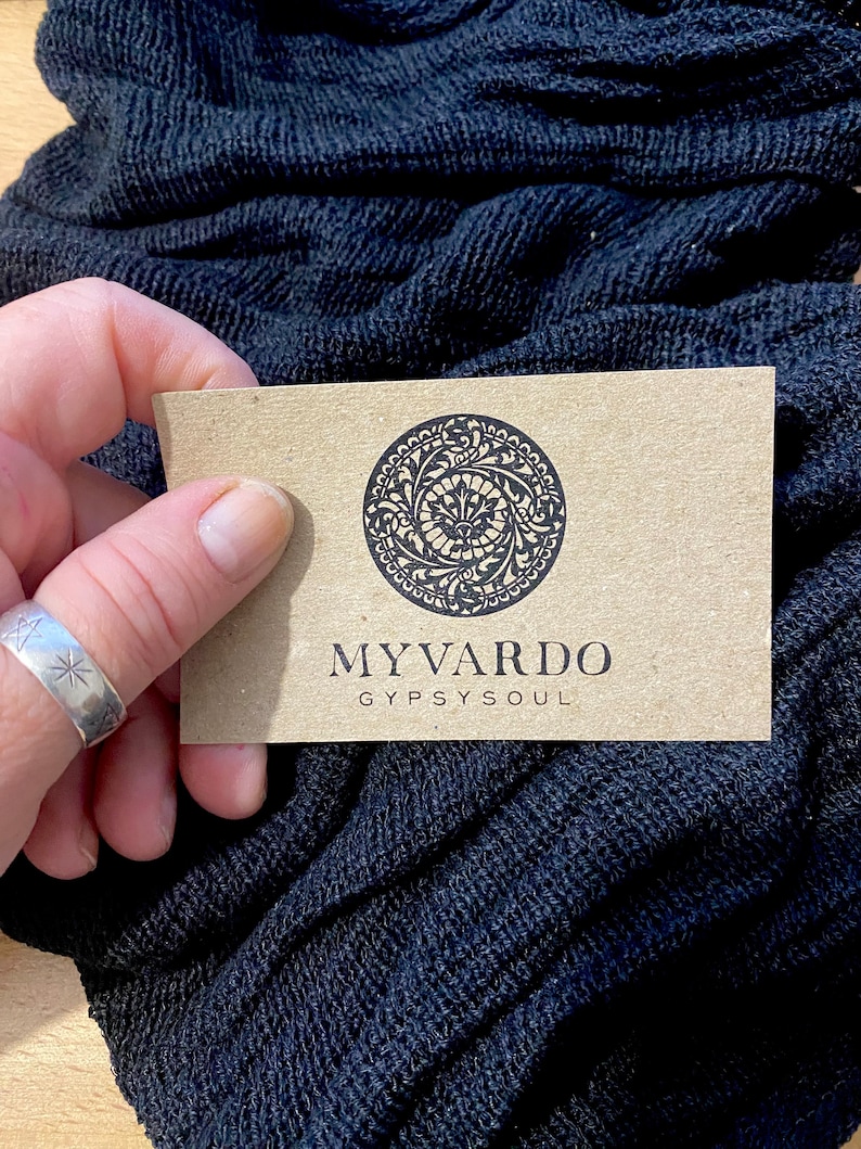 Op de afbeelding: Een lichtbruine visitekaart met de tekst "MYVARDO GYPSYSOUL" en een cirkelvormig ontwerp wordt boven een donkerblauwe, getextureerde stof gehouden. Een zilveren ring met sterontwerpen is zichtbaar. De stof lijkt een sjaal te zijn.