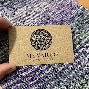 Op de afbeelding: Een close-up van een bruin label met de zwarte tekst "MYVARDO GYPSYSOUL". Het label heeft een cirkelvormig ontwerp. Het label wordt vastgehouden boven een gebreide stof met paarse, groene en witte strepen.