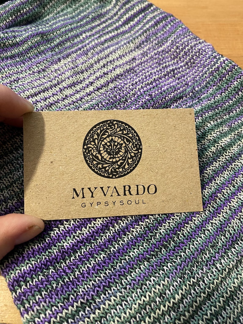 Op de afbeelding: Een close-up van een bruin label met de zwarte tekst "MYVARDO GYPSYSOUL". Het label heeft een cirkelvormig ontwerp. Het label wordt vastgehouden boven een gebreide stof met paarse, groene en witte strepen.