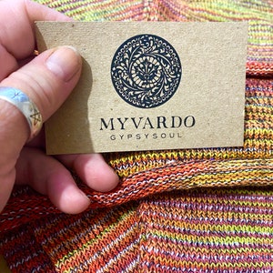 Op de afbeelding: Een close-up van een label met de zwarte tekst "MYVARDO GYPSYSOUL". Het label wordt boven een kleurrijke gebreide stof met oranje, gele en paarse tinten gehouden. Het label heeft een cirkelvormig ontwerp met een sierlijk patroon.