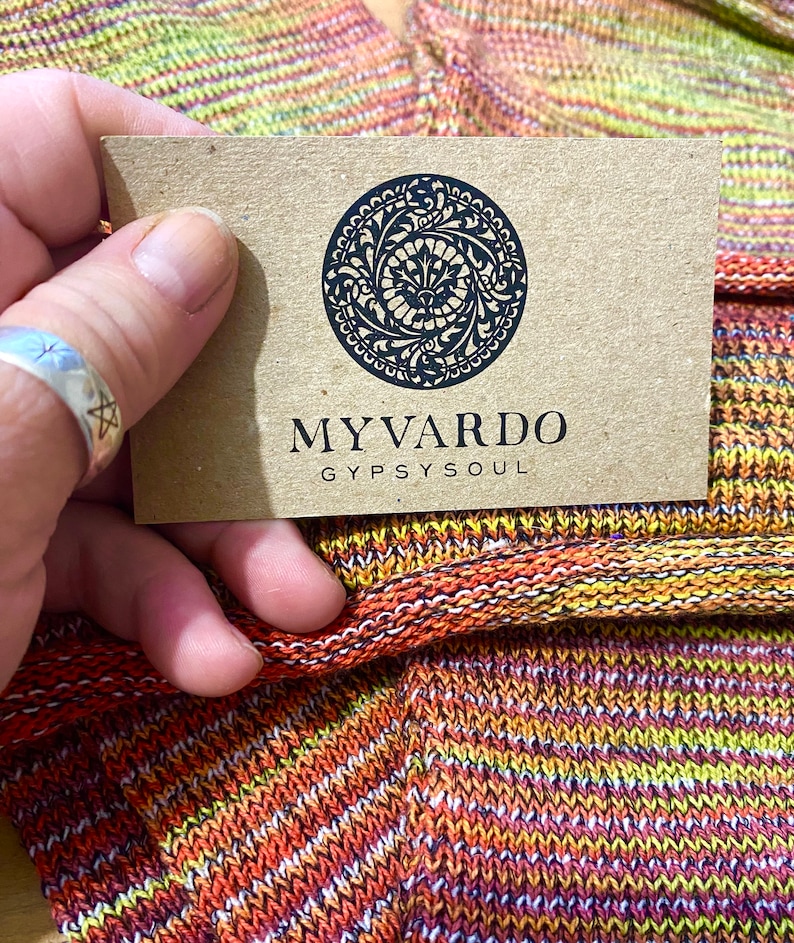 Op de afbeelding: Een close-up van een label met de zwarte tekst "MYVARDO GYPSYSOUL". Het label wordt boven een kleurrijke gebreide stof met oranje, gele en paarse tinten gehouden. Het label heeft een cirkelvormig ontwerp met een sierlijk patroon.