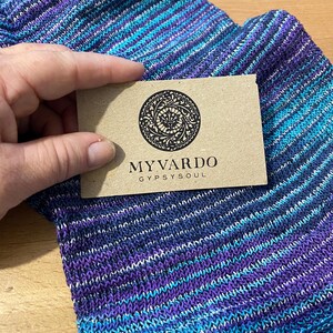 Op de afbeelding: Een hand houdt een bruin label met de tekst "MYVARDO GYPSYSOUL" boven een paar blauwe, paarse en wit gestreepte sokken. De sokken hebben een textuur gebreid patroon en een kleurengradi&euml;nt.