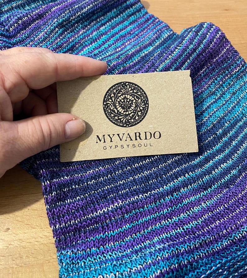 Op de afbeelding: Een hand houdt een bruin label met de tekst "MYVARDO GYPSYSOUL" boven een paar blauwe, paarse en wit gestreepte sokken. De sokken hebben een textuur gebreid patroon en een kleurengradi&euml;nt.