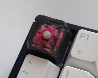 May include: A black and white keyboard with a custom keycap. The keycap is made of clear resin with pink and white swirls and a white round keycap stem. The keycap is labeled with the letter "F1".