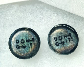 Puede incluir: Un par de pendientes redondos, negros e iridiscentes con el texto "DON'T QUIT" en letras blancas.