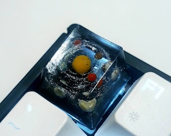 May include: A clear resin keycap with a galaxy design and a yellow sphere in the center. The keycap is on a white keyboard.