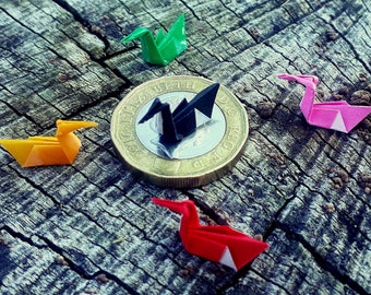 May include: Five origami paper cranes in different colors, including green, black, orange, pink, and red, are arranged around a British pound coin. The coin features the profile of Queen Elizabeth II and the inscription "2016 ELIZABETH II D.G. REG. F.D."