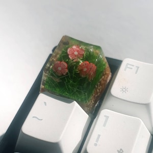 Könnte beinhalten: Eine transparente Harz-Tastenkappe mit einer Miniatur-Gartenszene mit grünem Gras und drei roten Blumen. Die Tastenkappe liegt auf einer weißen Tastatur.