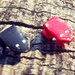 May include: Two small frog figurines, one red and one black, with white eyes, sitting on a wooden surface.
