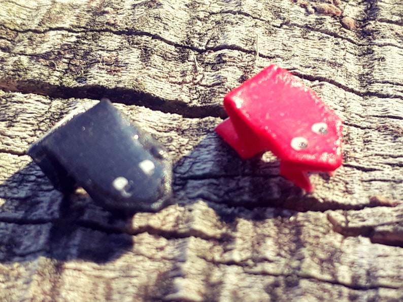May include: Two small frog figurines, one red and one black, with white eyes, sitting on a wooden surface.
