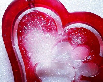 May include: Red heart-shaped resin ornament with a white paw print inside. The ornament is filled with glitter.