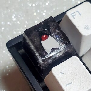 May include: A black and white keycap with a red and white design. The keycap has a glittery finish and is designed to fit a standard mechanical keyboard.