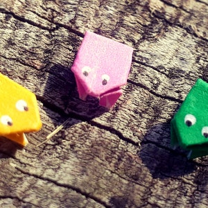 May include: Three origami frogs made of paper, one yellow, one pink, and one green, with white googly eyes. The frogs are sitting on a piece of wood.