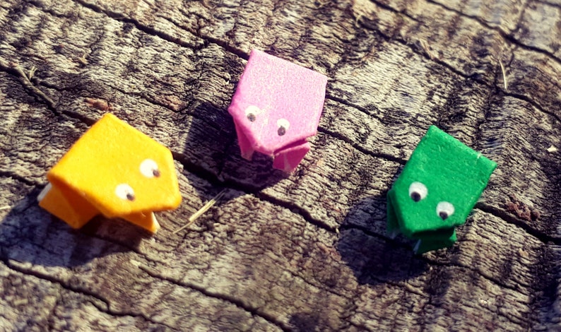 May include: Three origami frogs made of paper, one yellow, one pink, and one green, with white googly eyes. The frogs are sitting on a piece of wood.
