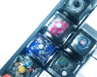 May include: A close-up of custom keyboard keycaps with a space theme. The keycaps are clear with colorful designs of planets, galaxies, and stars. The keycaps are square and sit on a black keyboard.
