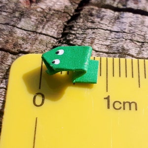 May include: A small green origami frog with white eyes, measuring approximately 1 centimeter in length, is placed on a yellow ruler.