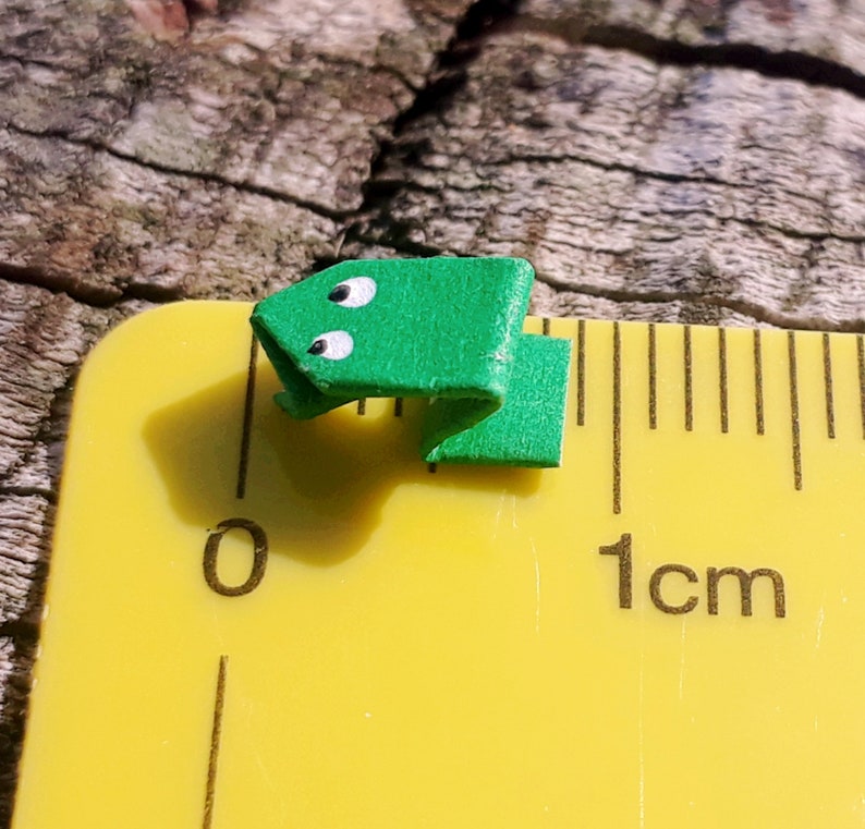 May include: A small green origami frog with white eyes, measuring approximately 1 centimeter in length, is placed on a yellow ruler.