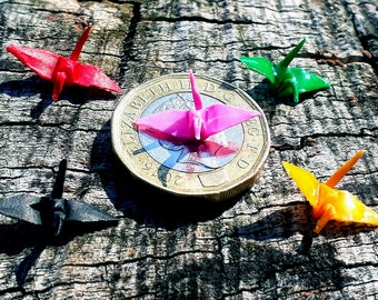 May include: Five origami paper cranes in different colors, including red, pink, green, black, and yellow, are arranged around a British one pound coin. The coin features the image of Queen Elizabeth II and the text "Elizabeth II D.G. REG. F.D. 2019."