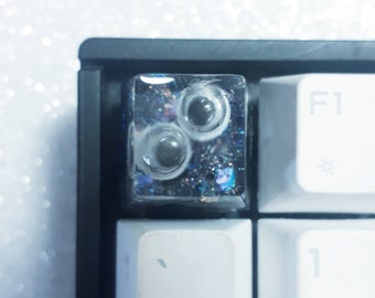 May include: A clear, square-shaped keycap with a glittery, iridescent finish. The keycap has three black, round, raised dots on the surface.