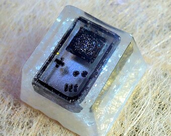 May include: A clear resin keycap featuring a black and white image of a Game Boy handheld video game console.