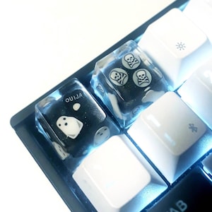 Puede incluir: Un teclado blanco con dos teclas personalizadas. Las teclas son transparentes y tienen diseños negros. Una tecla tiene un diseño de fantasma y la palabra "OUIJA" impresa en ella. La otra tecla tiene tres diseños de calaveras.