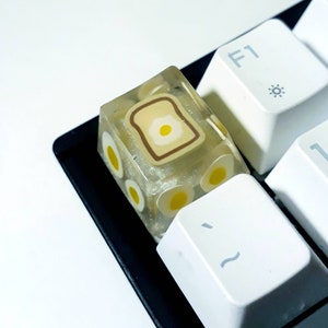 May include: A clear resin keycap with a brown and yellow design of a piece of toast with a fried egg on top. The keycap is on a white keyboard.