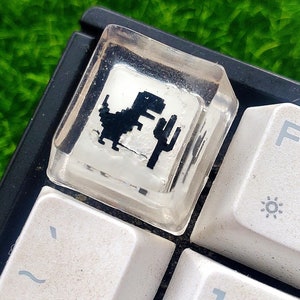 May include: A clear resin keycap with a black and white pixelated dinosaur graphic. The keycap is on a black keyboard with white keys.