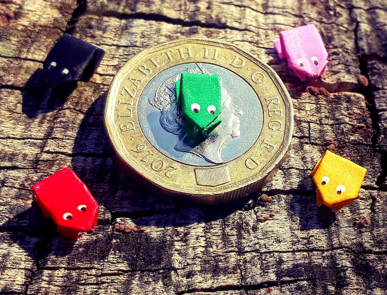 May include: Five origami frogs made of paper in different colors, including green, pink, black, red, and yellow, are arranged on a wooden surface. A gold British pound coin is in the center of the image.