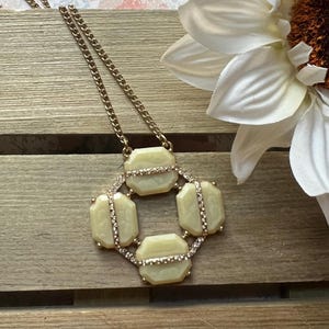 May include: A gold-toned necklace with a geometric pendant featuring pale yellow, octagonal stones. The stones are accented with a line of small, clear crystals. The necklace is displayed on a wooden surface, with a white flower in the background.