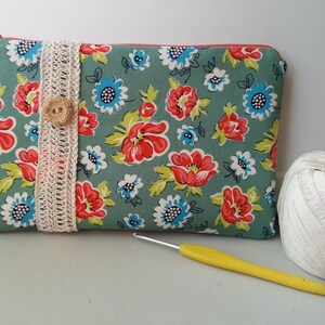 May include: A teal blue zippered pouch with a floral print featuring red, white, and blue flowers. The pouch has a white crocheted trim and a brown button.