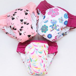 May include: Three cloth diapers with colorful patterns. One is pink with ballerina figures, another is white with a cat and floral design, and the third has a colorful swirl pattern. Each has a pink or burgundy trim.